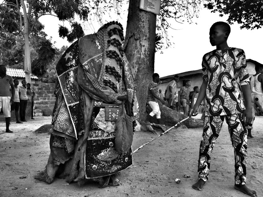 Un Egungun pr&ecirc;t &agrave; bondir et son gardien arm&eacute; d'un b&acirc;ton.